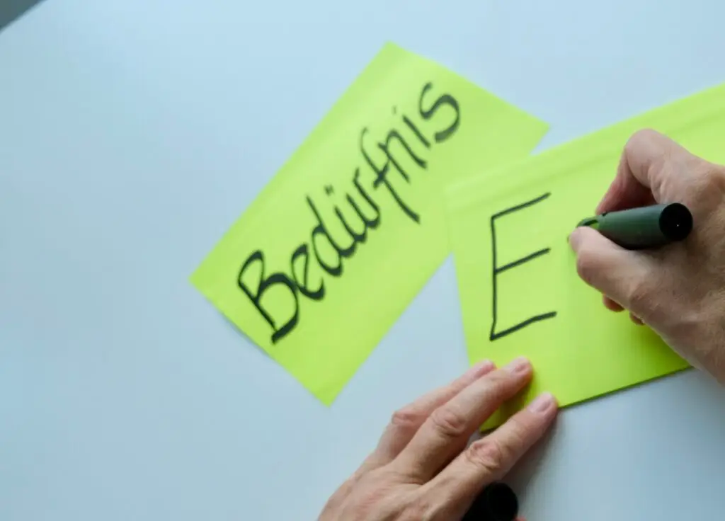 Eine Hand schreibt auf einen Aufkleber ein E und auf einem anderen steht bereits Bedürfnis